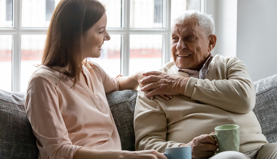 A woman talks to her senior father on the couch with coffee