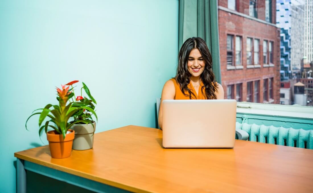 A young woman sits at a laptop working from a brightly colored home office